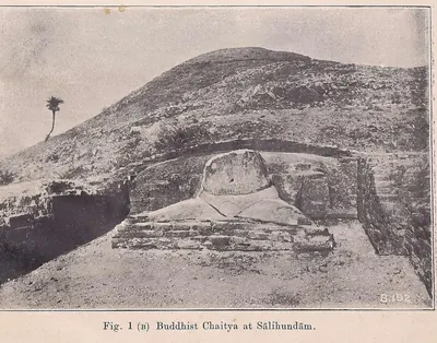 Photograph of an excavated brick shrine on a hillside, its curved back wall enclosing the broken lower half of a large seated plaster figure on a low brick platform, with a lone palm tree on the skyline.