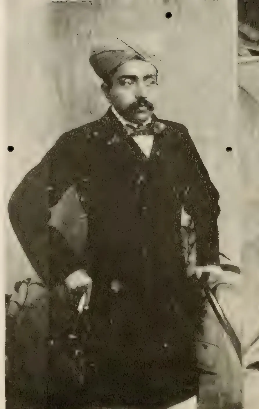 Old halftone photograph of a young man with a moustache, in a dark frock coat, bow tie and embroidered cap, standing with one hand on his hip beside a plant.