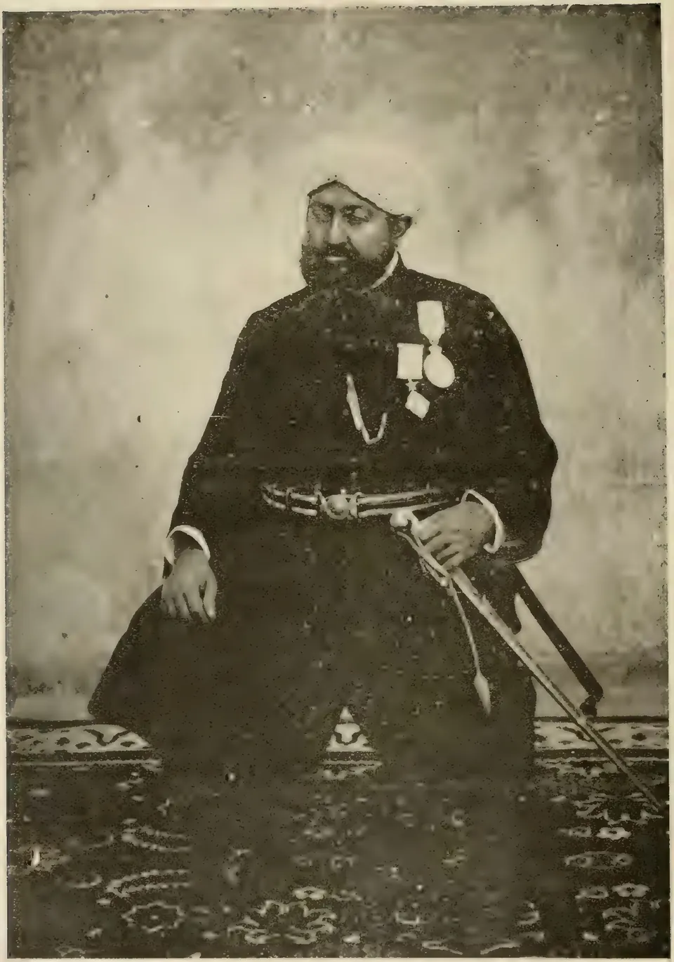 Old halftone photograph of a bearded man in a white turban and a long dark coat with two medals, seated with a sword across his knees on a patterned carpet before a painted backdrop.