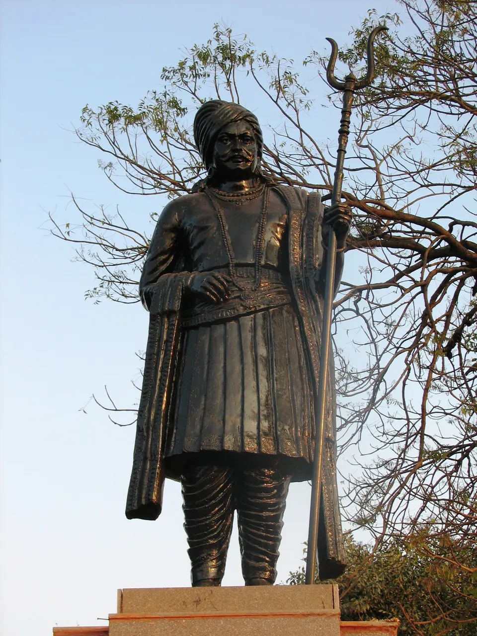 A bronze statue of a turbaned man in a long coat holding a trident-topped staff, on a stone pedestal against a blue sky and a leafless tree.