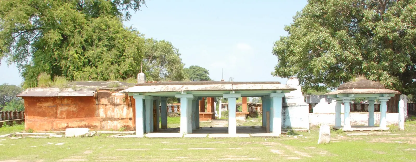 A low open pillared hall with a flat roof and smaller shrines on a grassy compound under large trees.
