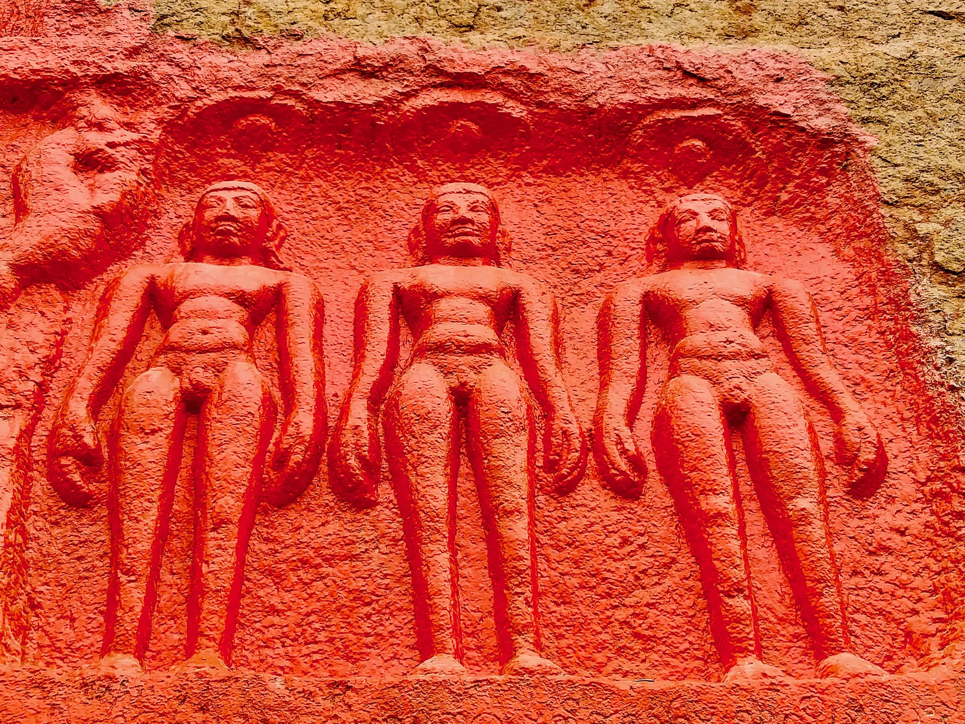 Panel of three standing nude Jaina figures carved side by side in a shallow niche on the rock, painted red.