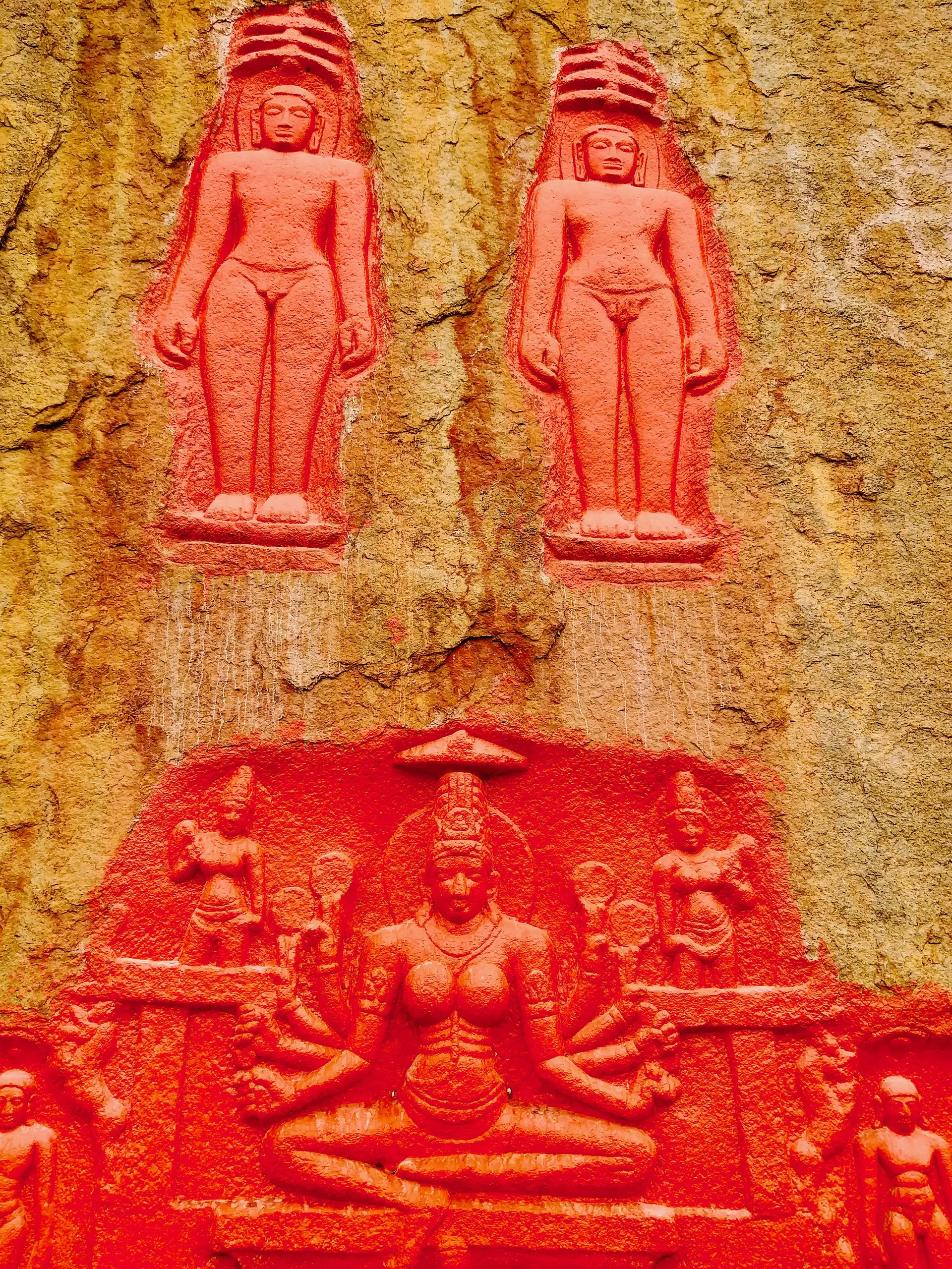 Rock face on the Bommala Gutta with two standing Jaina figures carved in shallow relief above a panel of a seated many-armed goddess with attendants, all painted bright red.