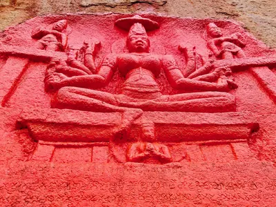 Close view of a relief of a seated goddess with eight arms holding attributes, flanked by small attendant figures and set in a framed niche, painted red; a band of inscribed letters is visible below.