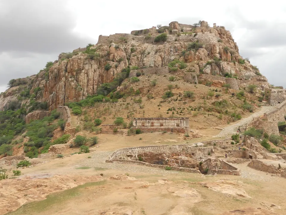 Colour photograph of a rocky hill with fortification walls winding up to the summit under a cloudy sky.