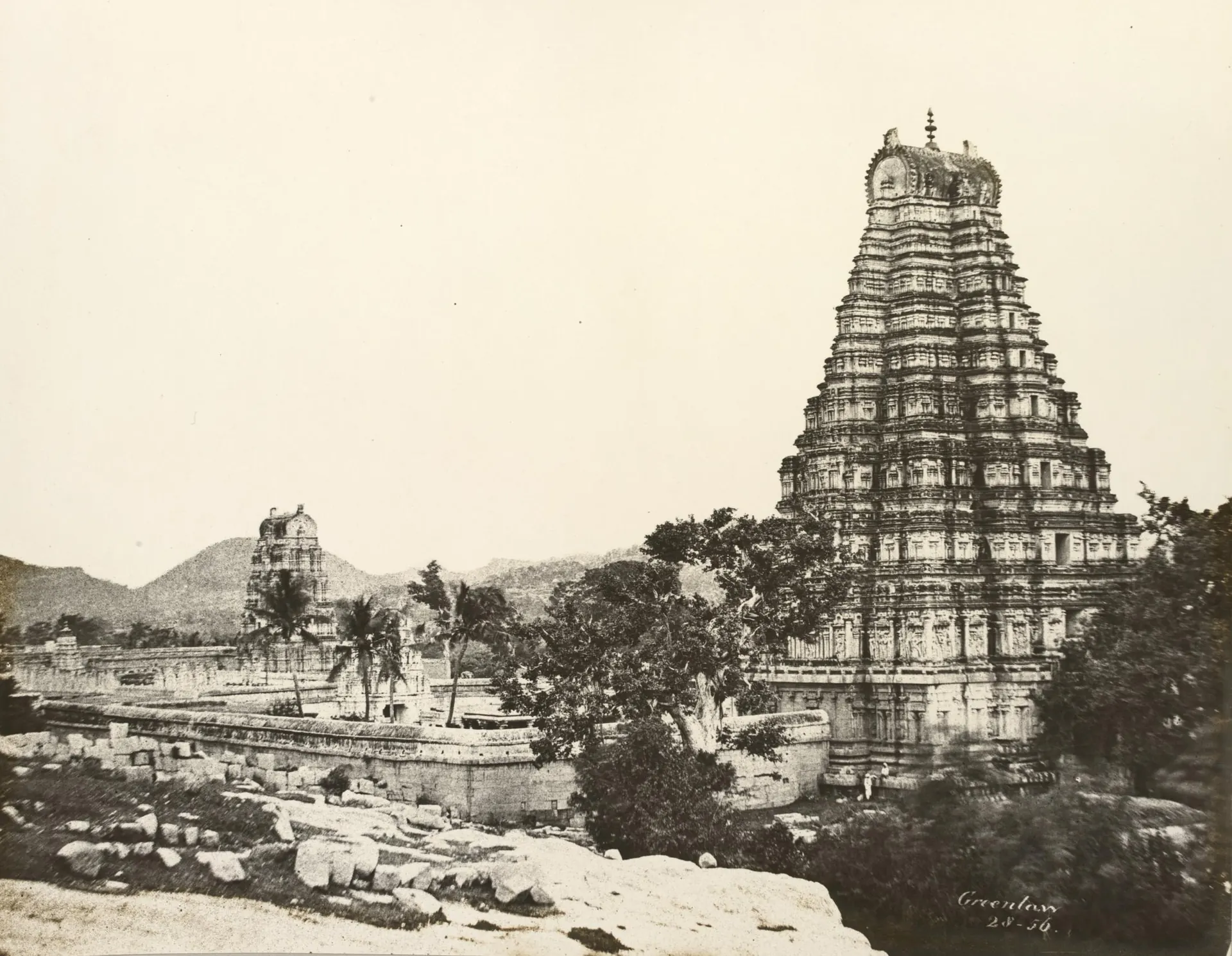 Sepia calotype of the tall gateway tower of the Virupaksha temple at Hampi rising above palms and ruined buildings, with rocky hills behind.