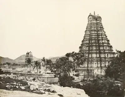 Sepia calotype of the tall gateway tower of the Virupaksha temple at Hampi rising above palms and ruined buildings, with rocky hills behind.
