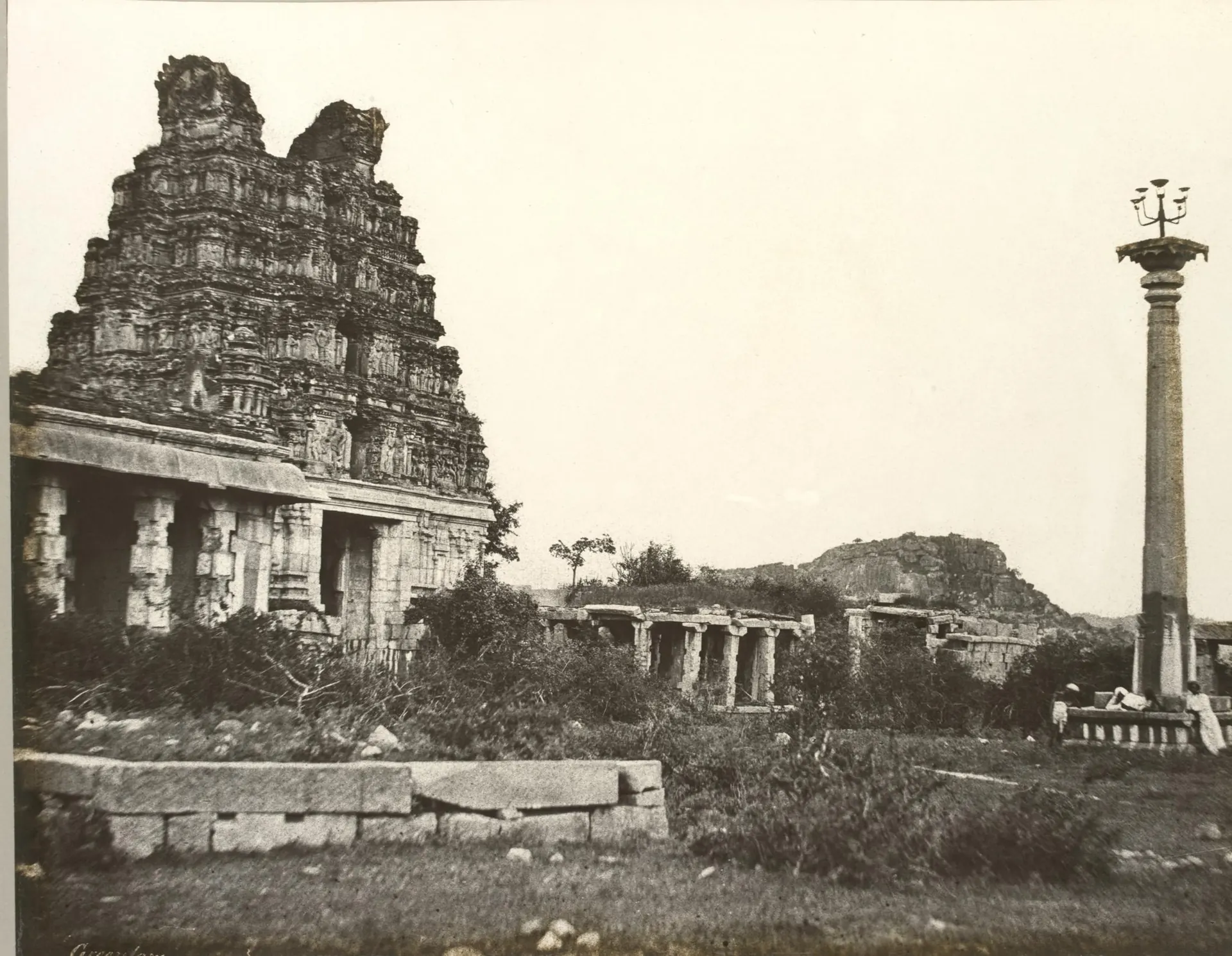 Sepia calotype of a ruined temple gateway tower beside pillared halls, with a tall slender lamp column standing in open ground at right.