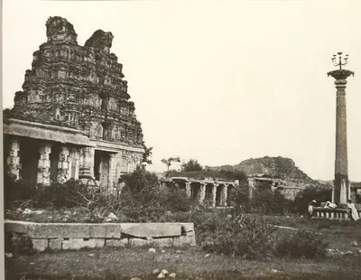 Sepia calotype of a ruined temple gateway tower beside pillared halls, with a tall slender lamp column standing in open ground at right.