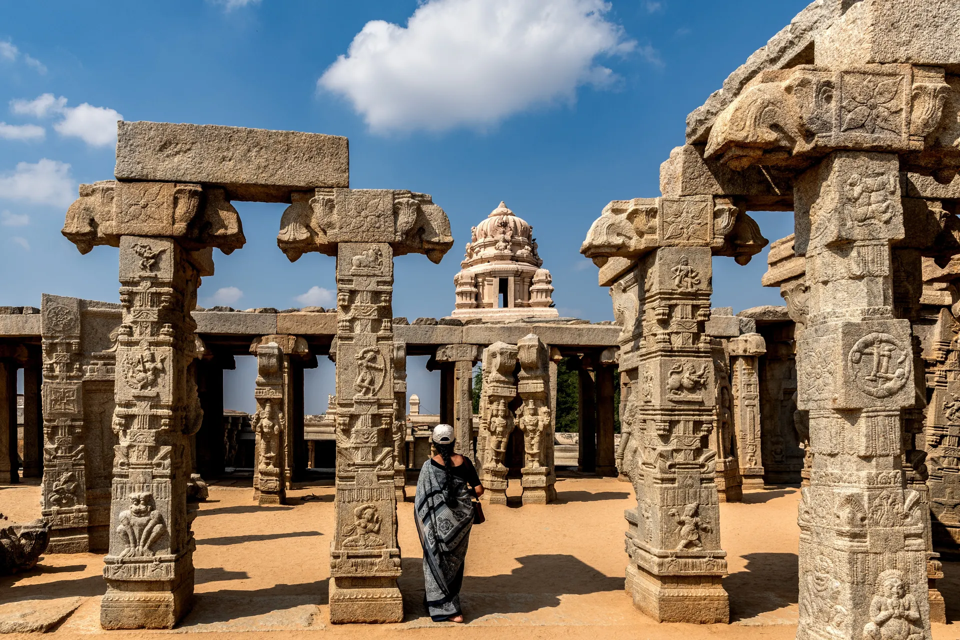 Colour photograph of carved granite pillars standing in the open court of the unfinished marriage hall at Lepakshi, a temple tower behind.