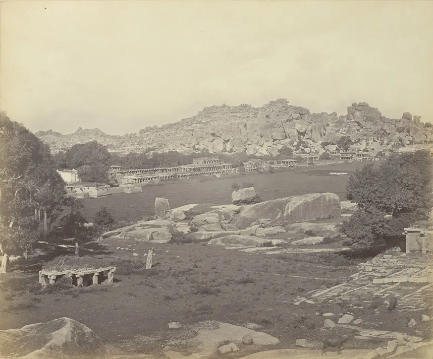 Albumen photograph of a long street lined with ruined stone pavilions and colonnades running towards boulder-strewn hills.
