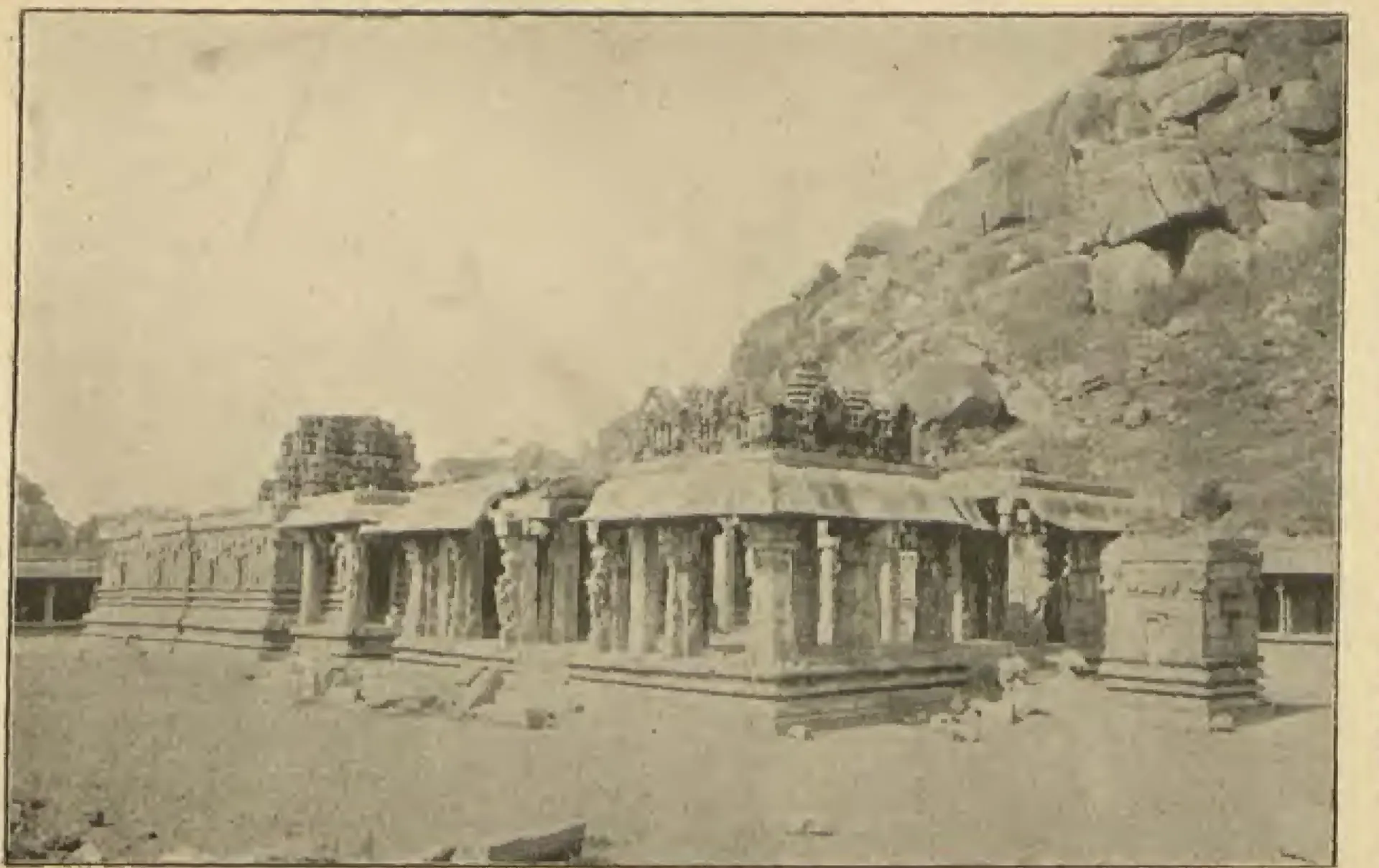 Photograph of the long pillared halls of the ruined temple of Achyuta Raya at Hampi, below a steep boulder-strewn hill. Printed sideways.