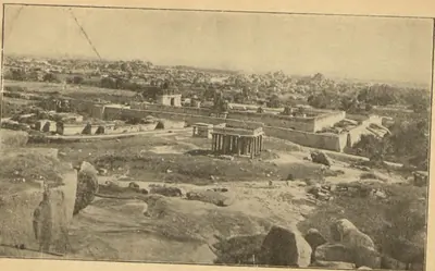 Photograph looking over the ruins of Vijayanagara from a hill: boulders in the foreground, a long pillared street and temple buildings in the middle distance and rocky hills beyond. Printed sideways.