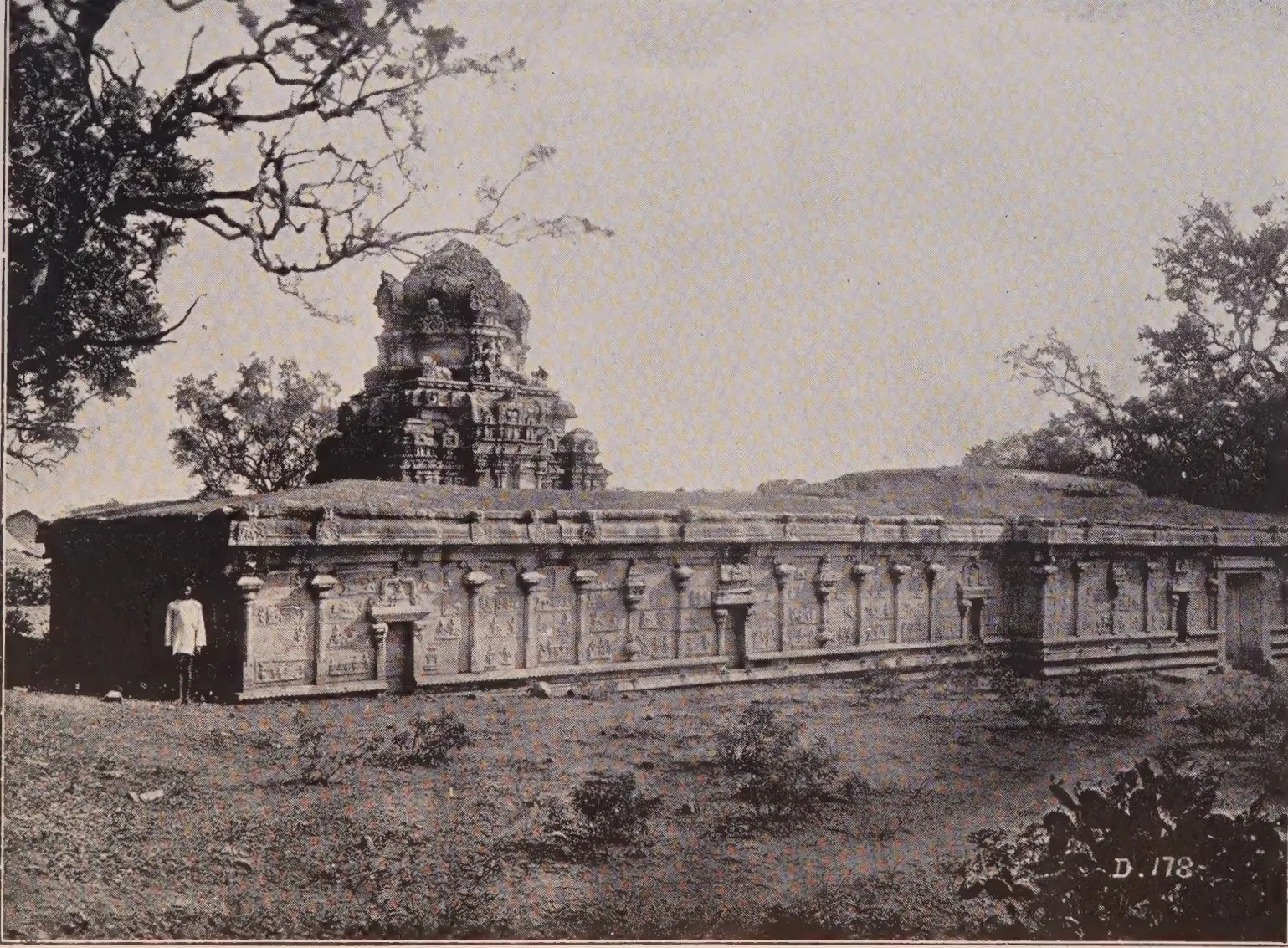 Photograph of a long flat-roofed stone temple with a carved basement and a pyramidal tower over the shrine, a man standing by its wall, trees behind.