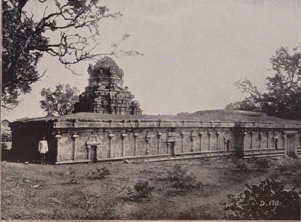 Photograph of a long flat-roofed stone temple with a carved basement and a pyramidal tower over the shrine, a man standing by its wall, trees behind.
