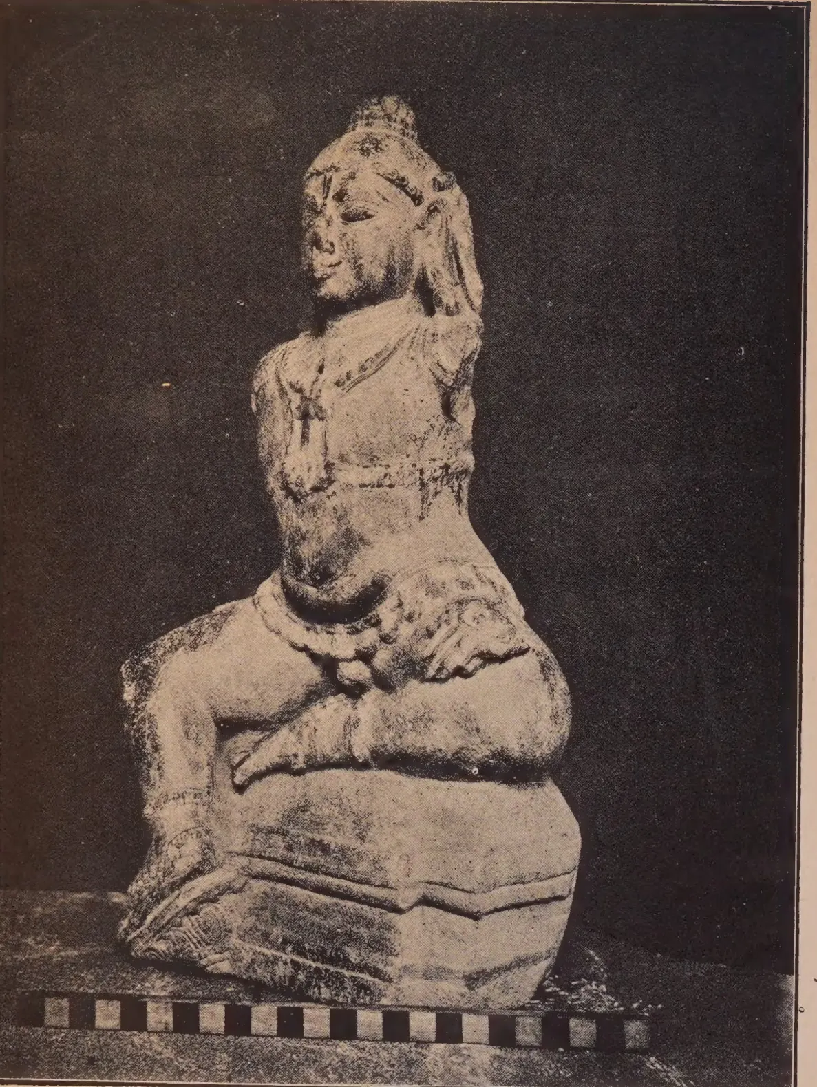 Photograph of a damaged stone statue of the child Krishna seated on a base, one knee raised, with a scale bar below, against a dark cloth.