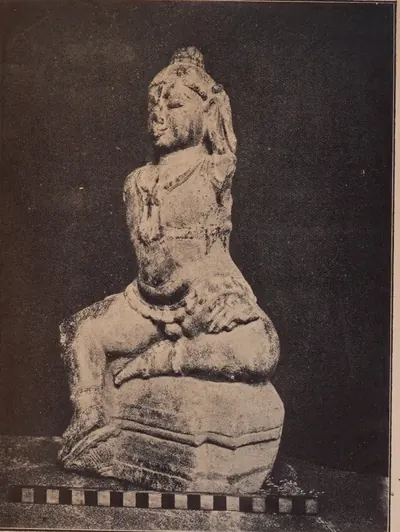 Photograph of a damaged stone statue of the child Krishna seated on a base, one knee raised, with a scale bar below, against a dark cloth.
