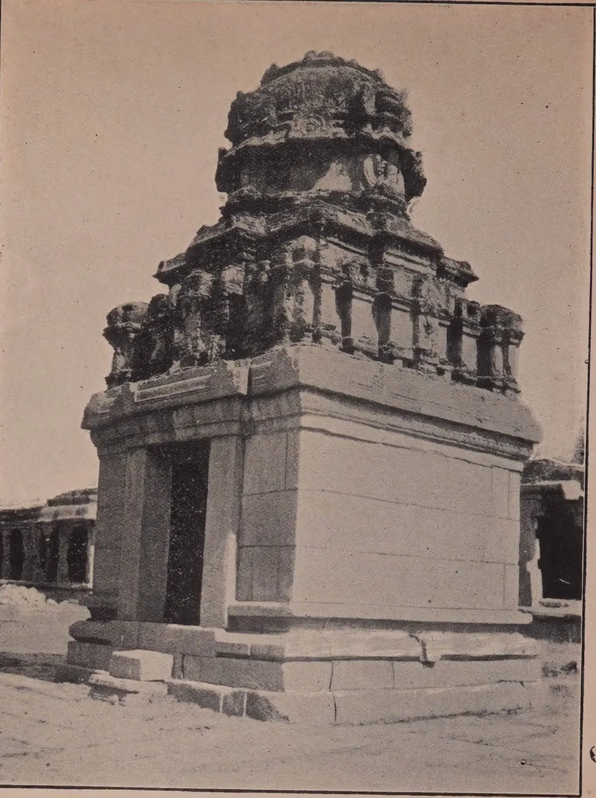 Photograph of a small square stone shrine with a plain doorway and a weathered tower, standing in a paved court.