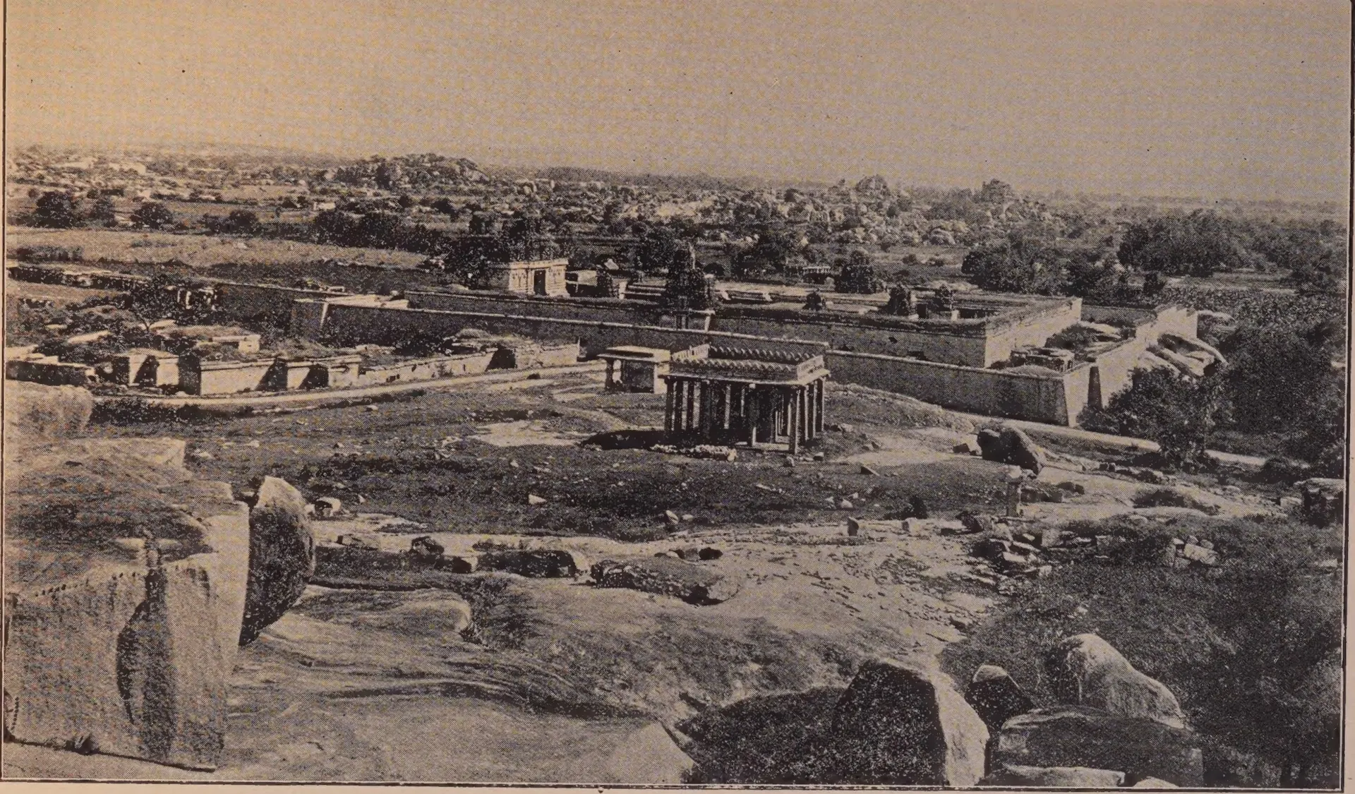 Photograph over boulders towards a walled temple enclosure with a pillared pavilion in front and the ruins of Vijayanagara beyond. Printed sideways.