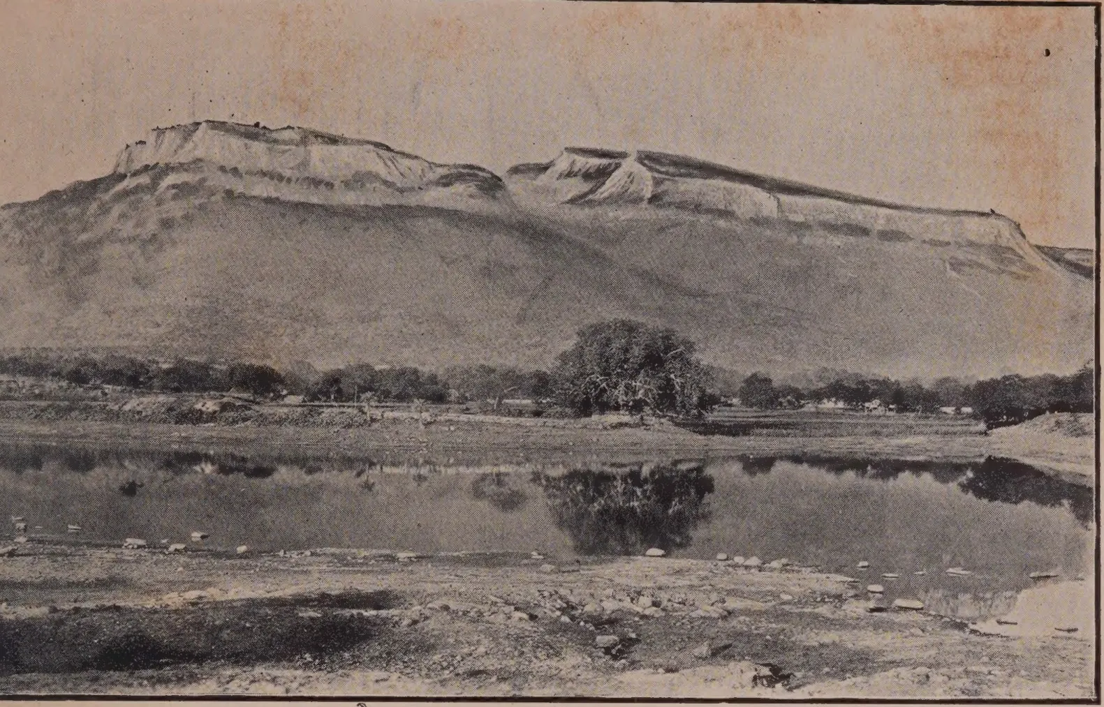 Photograph of the long flat-topped Udayagiri hill with steep cliffs rising behind a reach of still water and trees.