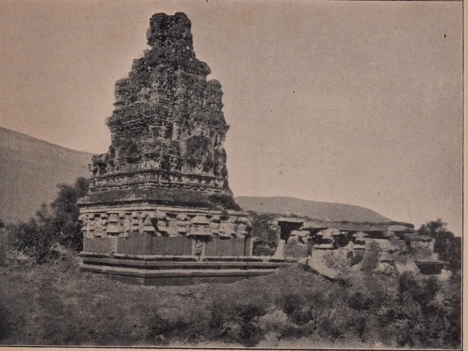 Photograph of a small stone temple with a tall weathered brick tower over its shrine, on a low rise among shrubs.