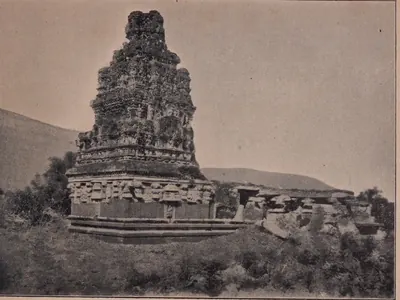 Photograph of a small stone temple with a tall weathered brick tower over its shrine, on a low rise among shrubs.