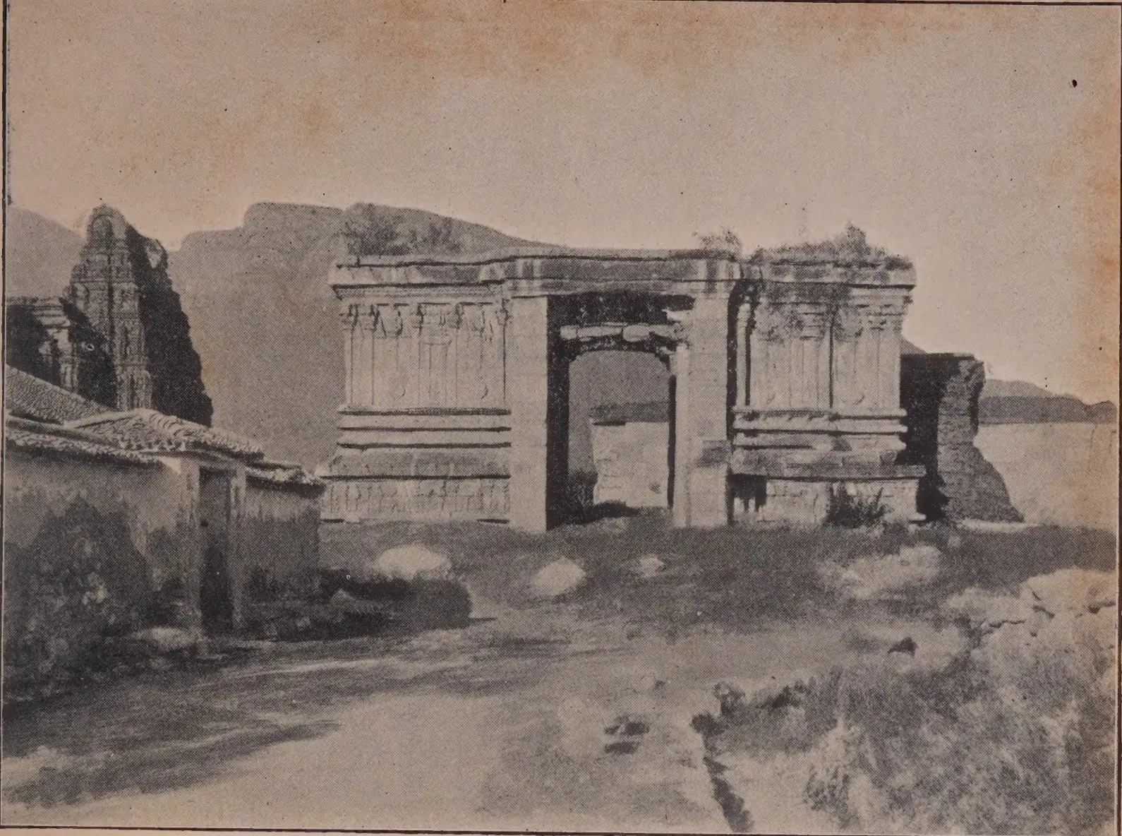 Photograph of the ruined stone base of a temple gateway with carved pilasters and an open doorway, a temple tower and a tiled house at left, a hill behind.