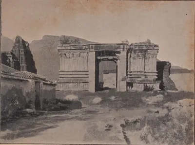 Photograph of the ruined stone base of a temple gateway with carved pilasters and an open doorway, a temple tower and a tiled house at left, a hill behind.