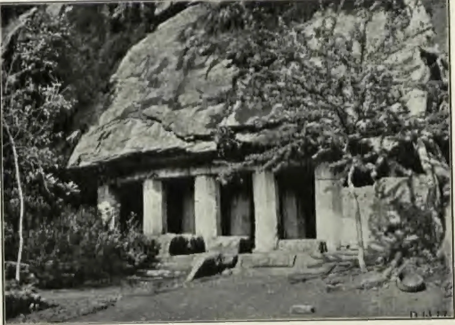 Old photograph of a low rock-cut cave temple at the foot of a boulder hill in Vijayawada: a row of square pillars forming the veranda, rock and a thorny tree above, scrub in front.