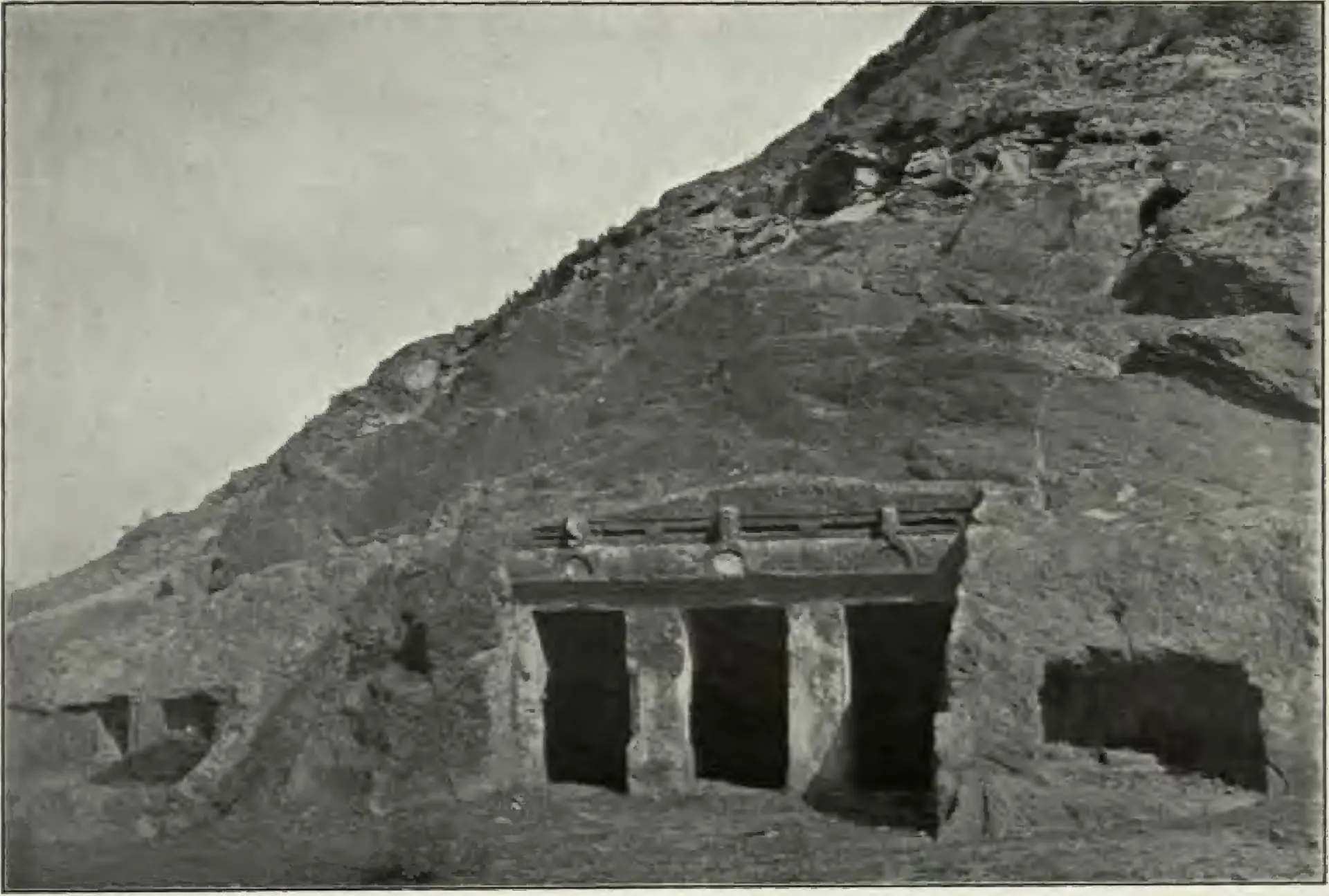 Old photograph of a small rock-cut shrine in a bare rocky hillside at Mogalrajapuram, Vijayawada: a facade with two square pillars and three dark openings under a projecting cornice, a second rough cutting to its right.