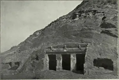 Old photograph of a small rock-cut shrine in a bare rocky hillside at Mogalrajapuram, Vijayawada: a facade with two square pillars and three dark openings under a projecting cornice, a second rough cutting to its right.