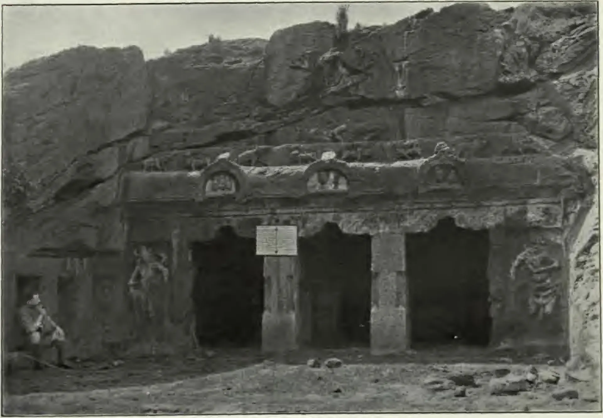 Old photograph of a larger rock-cut cave temple at Mogalrajapuram: a pillared facade with three openings below a cornice carved with arched gable-window motifs, a standing doorkeeper figure carved at the left, and a seated man at the far left.
