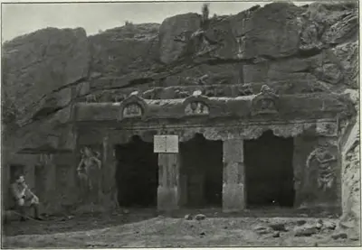 Old photograph of a larger rock-cut cave temple at Mogalrajapuram: a pillared facade with three openings below a cornice carved with arched gable-window motifs, a standing doorkeeper figure carved at the left, and a seated man at the far left.