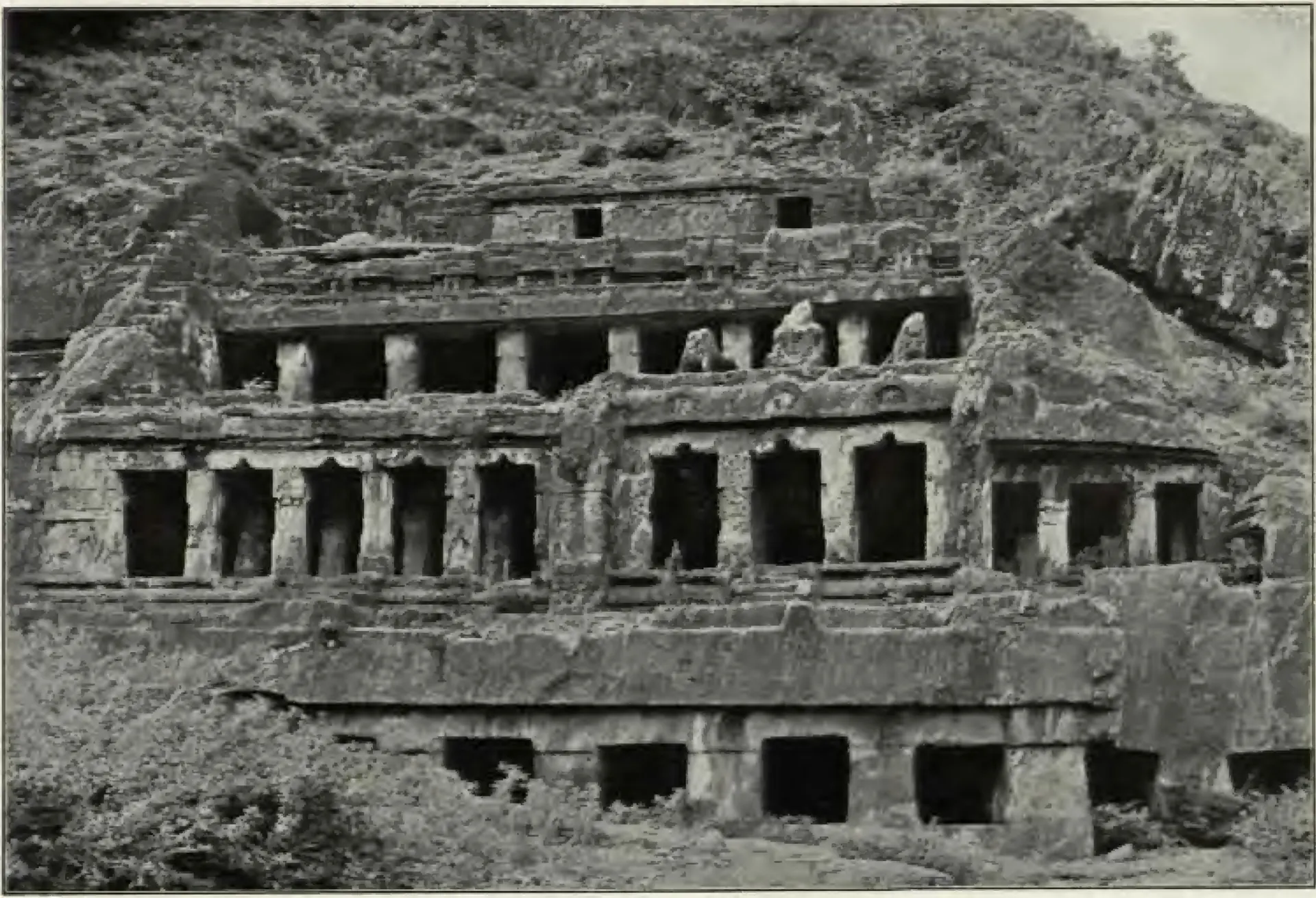Old photograph of the Undavalli rock-cut temple: the face of a hill carved into three tiers of pillared verandas above a lower row of dark openings, with vegetation on the rock above and scrub in front.