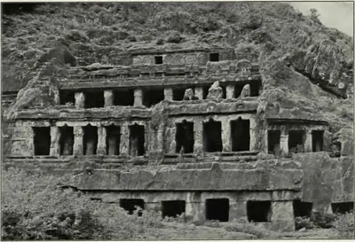 Old photograph of the Undavalli rock-cut temple: the face of a hill carved into three tiers of pillared verandas above a lower row of dark openings, with vegetation on the rock above and scrub in front.