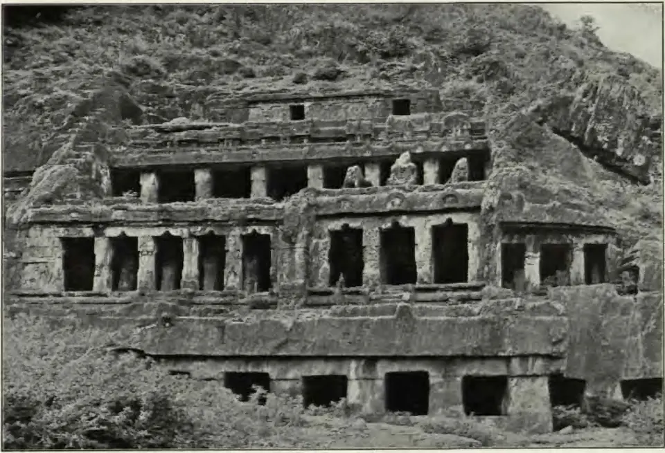 Old photograph of the Undavalli rock-cut temple: the face of a hill carved into three tiers of pillared verandas above a lower row of dark openings, with vegetation on the rock above and scrub in front.